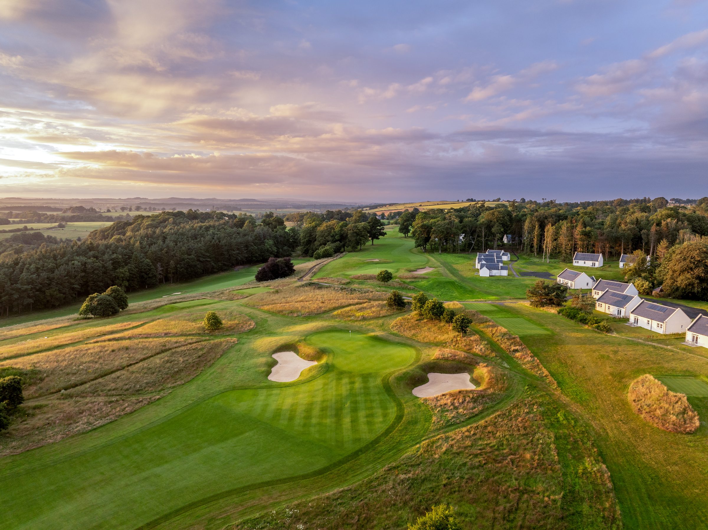 A green on the golf course at Schloss Roxburghe at dusk, guarded by two bunkers, with the Roxburghe Lodges beyond the trees